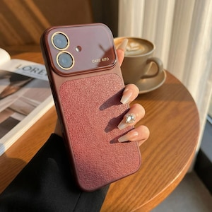 May include: A burgundy phone case with a textured lower section and a smooth upper section. The upper section has two camera cutouts with gold-colored rings and the text "CASE AITO". The case is held in a hand.