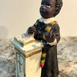 May include: A figurine of a young Black child in a black robe and white collar, standing behind a small white podium with a cross. The child has a colourful sash and is looking upwards. An open book rests on the podium.