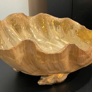 May include: A decorative, large, clam shell-shaped bowl. The bowl is a warm, golden color with a glossy finish. The edges have a wavy design, and the bowl rests on three small feet.