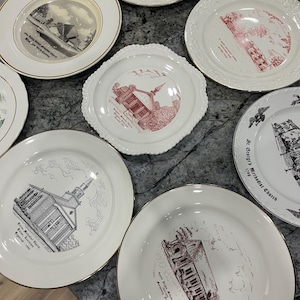 May include: A selection of vintage decorative plates, each with a different illustration of churches and historical buildings. The plates are white with gold rims and illustrations in black, red, and green. Some plates have text with the church name and location.
