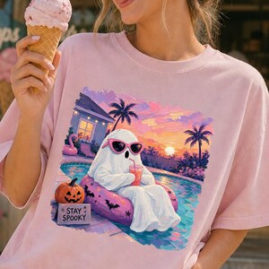 以下が含まれることがあります： ピンクのサングラスをかけた白い幽霊が、ピンクの浮き輪に乗ってプールに浮かび、飲み物を持っているグラフィックプリントが施された、ライトピンクの半袖Tシャツ。カボチャと「STAY SPOOKY」と書かれた看板が見える。