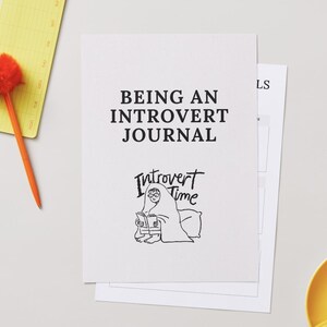 May include: A white journal with the text "BEING AN INTROVERT JOURNAL" and an illustration of a person reading under a blanket. An orange pen and notepad are to the left, and a yellow teacup is in the bottom right.