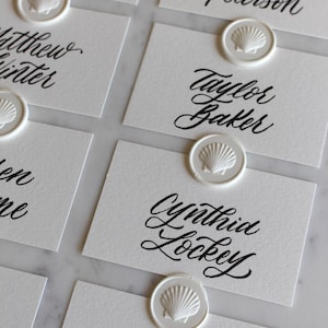 May include: White place cards with elegant black calligraphy names, each sealed with a white seashell wax stamp. The cards are arranged on a light-colored surface, perfect for a wedding or event.