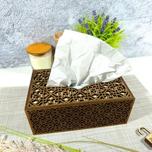 May include: A rectangular wooden tissue box with an intricate geometric cutout design. White tissues are pulled from the top opening. The box is brown, and the background includes a candle, a small vase, and a plant with purple flowers.