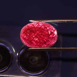 Peut inclure: Une pierre précieuse rouge foncé de forme ovale avec une surface à facettes et un motif interne en forme de grille visible. La pierre est maintenue entre les pointes métalliques d'une pince, révélant sa translucidité et sa couleur cramoisie.
