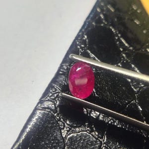 Puede incluir: Una gema de rubí de corte ovalado con un tono rojo intenso y superficie facetada, sostenida firmemente por unas pinzas de metal plateado. La gema muestra claridad interna y reflejos de luz en su superficie pulida y multifacetada.