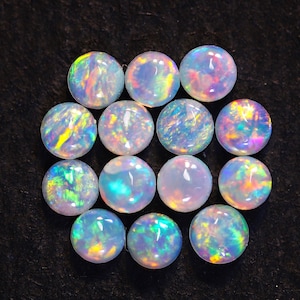 May include: A collection of twelve round, cabochon-cut opals arranged in a cluster. Each stone features a translucent white base with vibrant, multi-colored flashes of green, blue, yellow, and pink light reflecting from within the polished surfaces.
