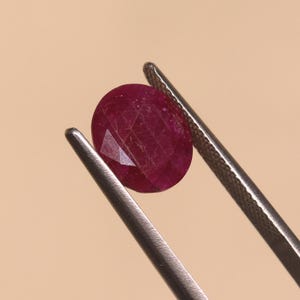 May include: A deep red, oval-shaped faceted gemstone held by a pair of silver-toned metal tweezers. The stone features a complex pattern of geometric facets that reflect light across its surface, showcasing a rich, saturated crimson color throughout.
