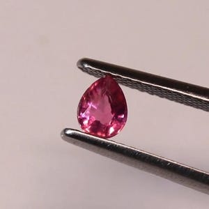 Puede incluir: Una gema rosa brillante en forma de pera es sostenida por unas pinzas plateadas. La gema tiene una superficie pulida y un color profundo y saturado. Las pinzas están enfocadas sobre un fondo suave y neutro.
