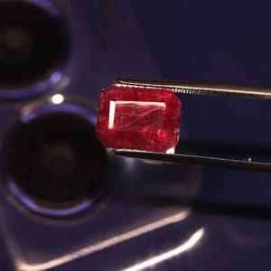 Puede incluir: Una piedra preciosa roja rectangular y facetada sujeta por unas pinzas metálicas. La piedra muestra un tono carmesí intenso con inclusiones internas visibles y bordes geométricos afilados. La superficie refleja la luz con claridad.