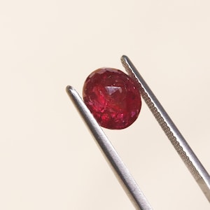Puede incluir: Una piedra preciosa de color rojo intenso, de forma ovalada y facetada, sostenida por unas pinzas de metal plateado. La piedra muestra una superficie texturizada con múltiples facetas que refleja la luz, exhibiendo un tono carmesí.