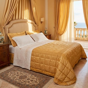 May include: A luxurious bedroom with a large bed featuring a gold quilted comforter and white linens. The room has gold curtains, a patterned rug, and a view of the ocean. Decorative lamps and a headboard complete the elegant setting.