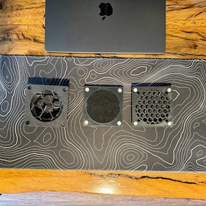 May include: A black laptop sits above a black and white topographic map mousepad. Three black square fan covers with different geometric designs are on the mousepad. The desk is made of wood.