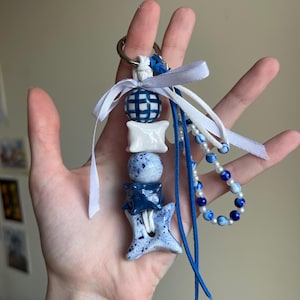 Peut inclure: Un porte-clés artisanal avec des éléments en céramique et perles bleues et blanches. Il comprend une sphère à carreaux, une perle en forme de coussin et un charm en forme d'étoile, reliés par un anneau métallique et des rubans.