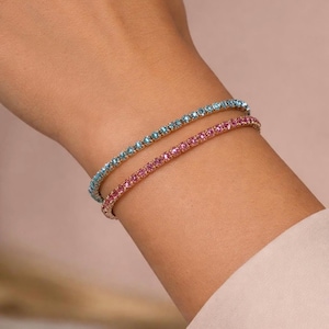 May include: Two delicate bracelets, one light blue and one pink, are adorned with small, sparkling stones. The bracelets are worn on a wrist, showcasing their elegant design and vibrant colours. The jewellery adds a touch of glamour.