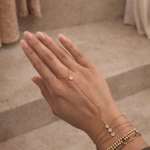 May include: Gold-toned hand chain bracelet with three clear, round gemstones and a delicate chain that connects to a finger ring. Several gold bracelets with various designs are stacked on the wrist.