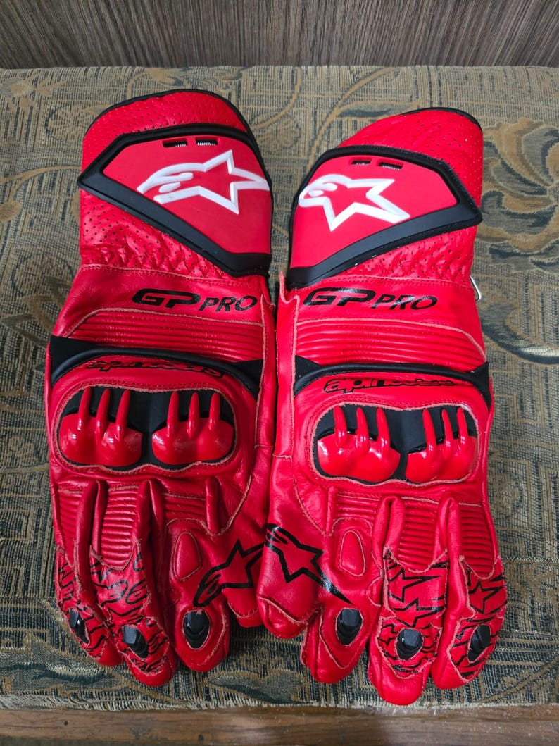 May include: Pair of red and black motorcycle gloves with the text "GP PRO" and a white star logo. The gloves have protective padding on the knuckles and fingers, and black accents. The gloves are made of leather.