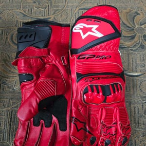 May include: A pair of red leather motorcycle gloves with black accents. The gloves feature the brand name "GP PRO" and a white star logo. The gloves have reinforced knuckles and finger protection, designed for safety and performance.