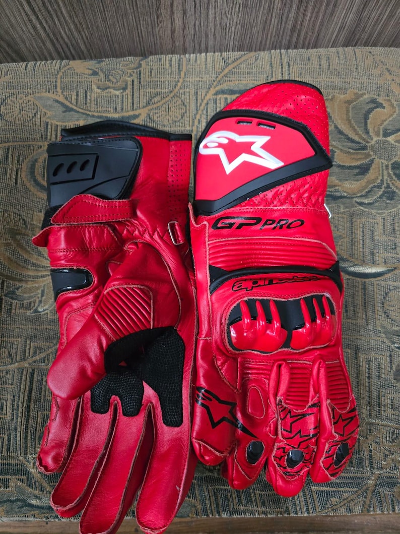 May include: A pair of red leather motorcycle gloves with black accents. The gloves feature the brand name "GP PRO" and a white star logo. The gloves have reinforced knuckles and finger protection, designed for safety and performance.