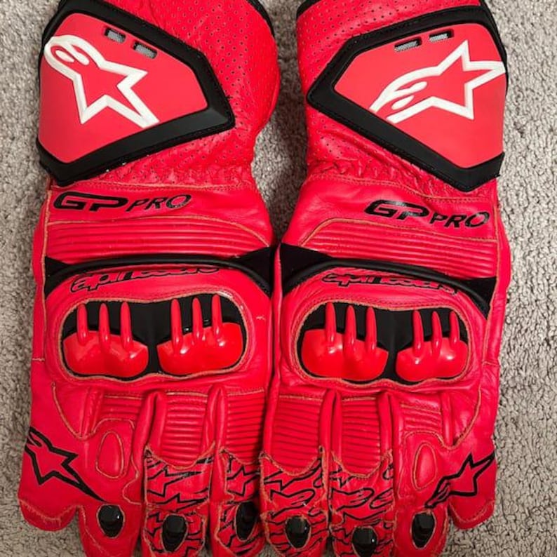 May include: Pair of bright red motorcycle gloves with black accents. The gloves feature the brand's logo, a white star within a shield, and the text "GP PRO". The gloves have protective padding on the knuckles and fingers.