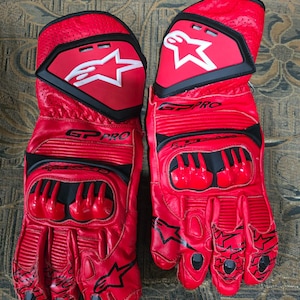 May include: A pair of red leather motorcycle gloves with black accents. The gloves feature the text "GP PRO" and a white star logo on the back of the hand. The gloves have protective padding on the knuckles and fingers.