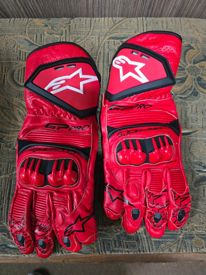 May include: A pair of red leather motorcycle gloves with black accents. The gloves feature the text "GP PRO" and a white star logo on the back of the hand. The gloves have protective padding on the knuckles and fingers.