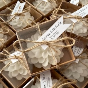 May include: Close-up of white, flower-shaped candles in clear boxes, tied with twine. Each box has a small tag with text. The candles are arranged in a close-up shot, creating a visually appealing display. The overall theme is wedding favors or event gifts.