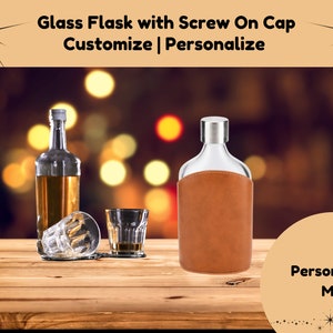 May include: A glass flask with a screw-on cap and a brown leather cover. The flask is sitting on a wooden table with a blurry background of lights. The text "Personalize Me!" is visible in the lower right corner of the image.