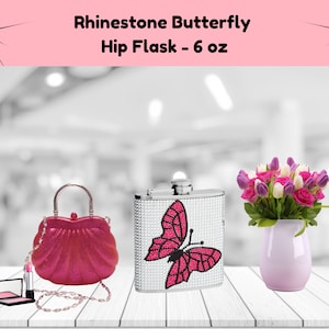 May include: A rhinestone butterfly hip flask with a pink butterfly design. The flask is 6 ounces and is sitting on a white wooden surface. There is a pink purse, a pink lipstick, and a pink blush compact next to the flask. There is a bouquet of pink and purple tulips in a light purple vase.