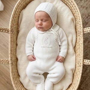 May include: A sleeping infant wearing a white long-sleeved knitted romper with footies and a matching bonnet. The romper features a central oval emblem with an embroidered letter E, a rounded collar, and cable knit detailing on the chest.