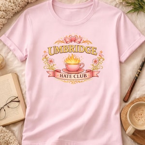 以下が含まれることがあります： 袖をまくった薄いピンク色のTシャツ。Tシャツには、炎の入ったティーカップの上に「Umbridge」の文字、バナーに「Hate Club」の文字が描かれたグラフィックがプリントされています。デザインは花の要素で囲まれています。