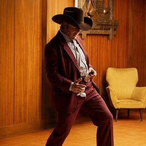 May include: A dark maroon suit consisting of a blazer and matching trousers, paired with a light-colored pinstriped button-down shirt, a dark brown cowboy hat, and a bolo tie with a prominent metallic belt buckle accessory at the waist.