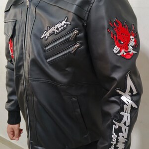May include: A black faux leather jacket featuring a grey hooded collar, a front zipper, and multiple pockets. The jacket is decorated with embroidered Cyberpunk 2077 logos on the chest and sleeve, along with a red and white oni mask patch.