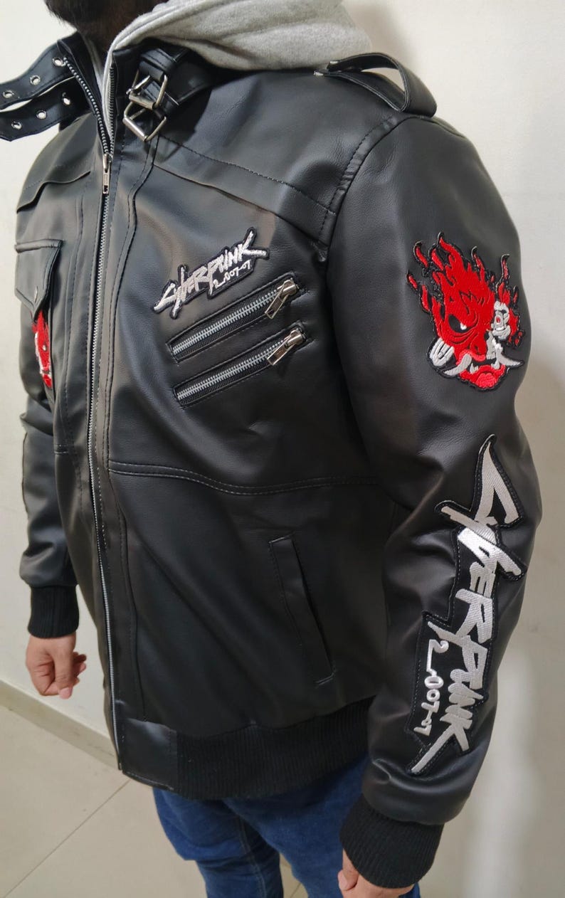 May include: A black faux leather jacket featuring a grey hooded collar, a front zipper, and multiple pockets. The jacket is decorated with embroidered Cyberpunk 2077 logos on the chest and sleeve, along with a red and white oni mask patch.