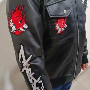 May include: A black faux leather jacket featuring a red and white embroidered samurai mask patch on the upper sleeve and chest pocket, with a large silver and black embroidered logo patch running down the lower sleeve and a front zipper closure.