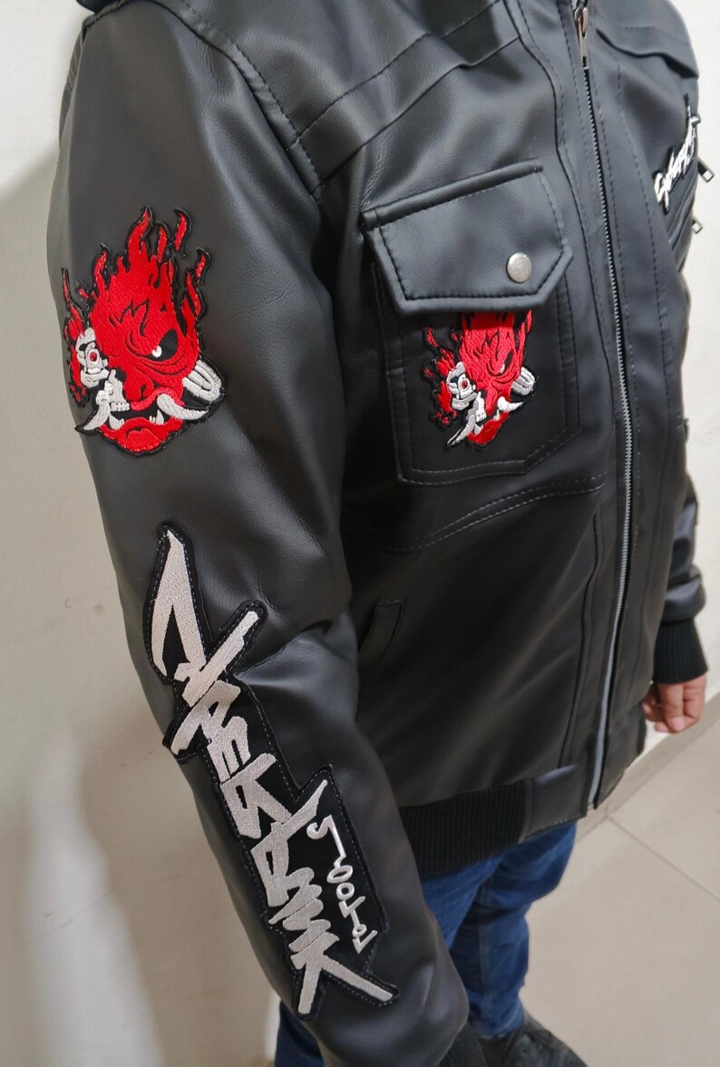 May include: A black faux leather jacket featuring a red and white embroidered samurai mask patch on the upper sleeve and chest pocket, with a large silver and black embroidered logo patch running down the lower sleeve and a front zipper closure.