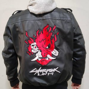 May include: A black faux leather bomber jacket featuring a large embroidered red and white oni mask graphic on the back. Below the mask, the text Cyberpunk 2077 is embroidered in white. The jacket has black ribbed cuffs and a light gray hooded sweatshirt underneath.
