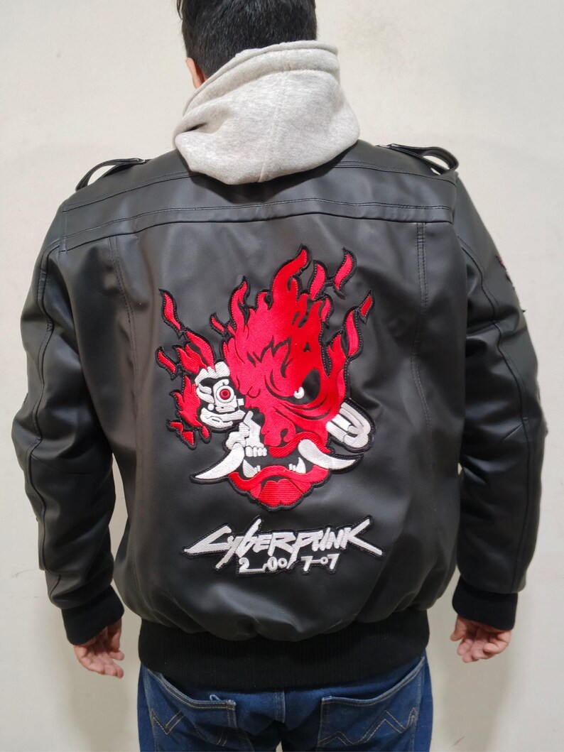 May include: A black faux leather bomber jacket featuring a large embroidered red and white oni mask graphic on the back. Below the mask, the text Cyberpunk 2077 is embroidered in white. The jacket has black ribbed cuffs and a light gray hooded sweatshirt underneath.