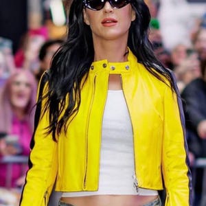 May include: A bright yellow cropped leather jacket with a high collar, silver-tone metal zipper, and black side panels. The jacket is worn over a plain white tank top and paired with blue denim jeans. The person is wearing dark oval sunglasses.