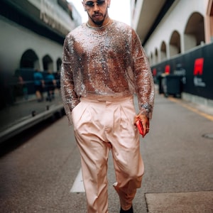 May include: A long-sleeved, semi-sheer top covered in shimmering silver sequins paired with light pink, high-waisted trousers featuring front pleats. The outfit is completed with dark shoes, a silver chain necklace, and tinted sunglasses.