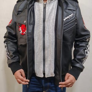 May include: A black faux leather jacket featuring a layered grey hooded insert, a stand-up collar with straps, and zippered pockets. The jacket displays a red oni mask patch on the chest, white text on the sleeves, and the word Cyberpunk in white.
