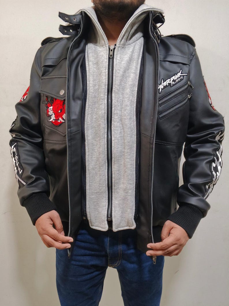 May include: A black faux leather jacket featuring a layered grey hooded insert, a stand-up collar with straps, and zippered pockets. The jacket displays a red oni mask patch on the chest, white text on the sleeves, and the word Cyberpunk in white.