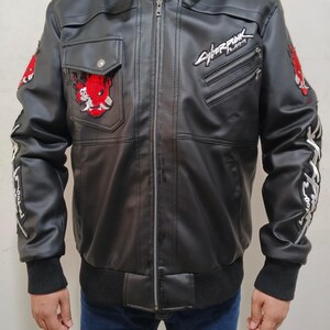 May include: A black faux leather jacket featuring a central zipper, a high collar with buckle straps, and ribbed cuffs. The chest displays a red oni mask patch and the Cyberpunk 2077 logo. Additional red patches and white text adorn the sleeves.
