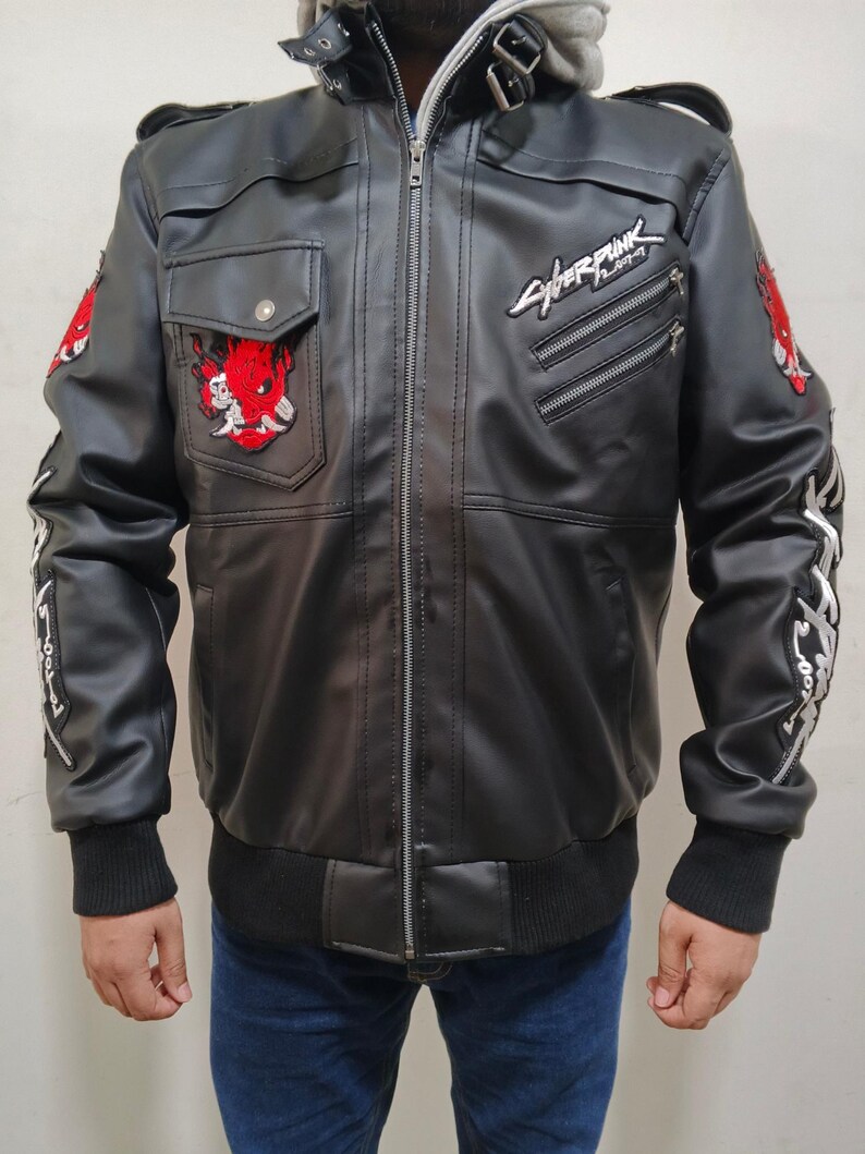 May include: A black faux leather jacket featuring a central zipper, a high collar with buckle straps, and ribbed cuffs. The chest displays a red oni mask patch and the Cyberpunk 2077 logo. Additional red patches and white text adorn the sleeves.