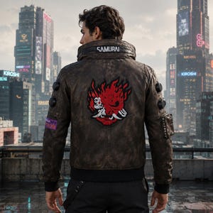 May include: A brown leather jacket featuring a high collar with the text SAMURAI printed on the back. The jacket displays a large red and black graphic of a stylized demon mask on the center back, along with circular black accents on the sleeves.
