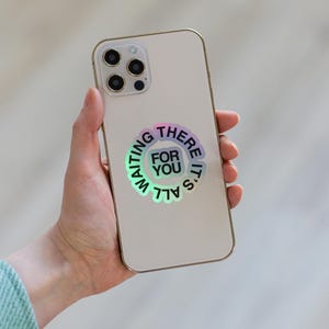 Può includere: Uno smartphone di colore chiaro con un adesivo olografico circolare sul retro. L'adesivo mostra il testo IT'S ALL WAITING THERE FOR YOU disposto in cerchio, con le parole FOR YOU centrate all'interno di un blocco di forma quadrata.