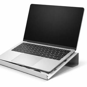 Könnte beinhalten: Ein silberner Laptop mit schwarzem Bildschirm und Tastatur steht auf einem silbernen Laptop-Ständer. Der Ständer neigt den Laptop und bietet eine ergonomische Einrichtung. Der Hintergrund ist weiß.