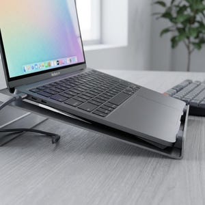 May include: A dark gray metal laptop stand holding a space gray laptop at an angled position. The laptop screen displays a colorful gradient background and a dock with various application icons. A gray keyboard and mouse sit to the right side.