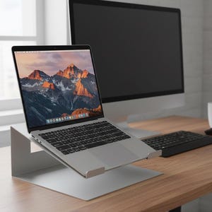 Peut inclure: Un ordinateur portable argenté sur un support argenté, affichant un paysage de montagne. Un clavier noir, un écran d'ordinateur, une souris et une tasse blanche sont sur un bureau en bois.