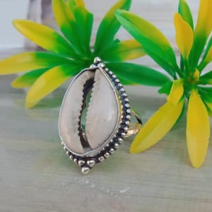 Könnte beinhalten: Silberfarbener Ring mit tropfenförmigem Design und natürlicher Kaurischale in der Mitte. Der Ring hat einen Perlenrand und ist vor einem Hintergrund aus grünen und gelben Kunstblättern platziert.